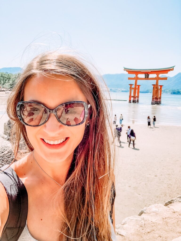 Sydney Travel Blogger, Bec Bowman on a solo trip to Japan, stands in front of the floating torii gate on Miyajima Island in Japan. The sky is blue and the sun is shining. The torii gate is red and stands in the distance at low tide.