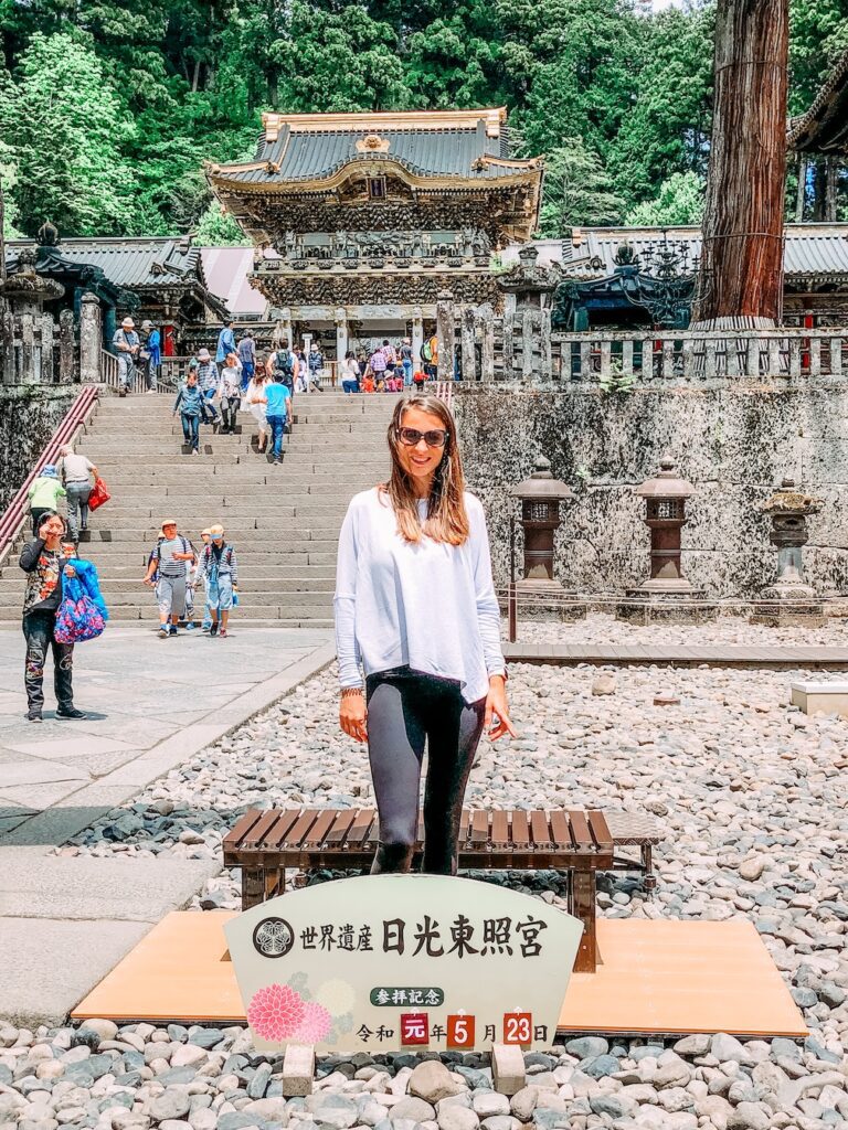 Bec standing outside the Nikko Toshogu Shrine in Japan. Known for its intricate carvings unlike any other in Japan. It is a sunny day and Bec is wearing sunglasses, a long sleeve grey top and black tights. There are crowds in the background.