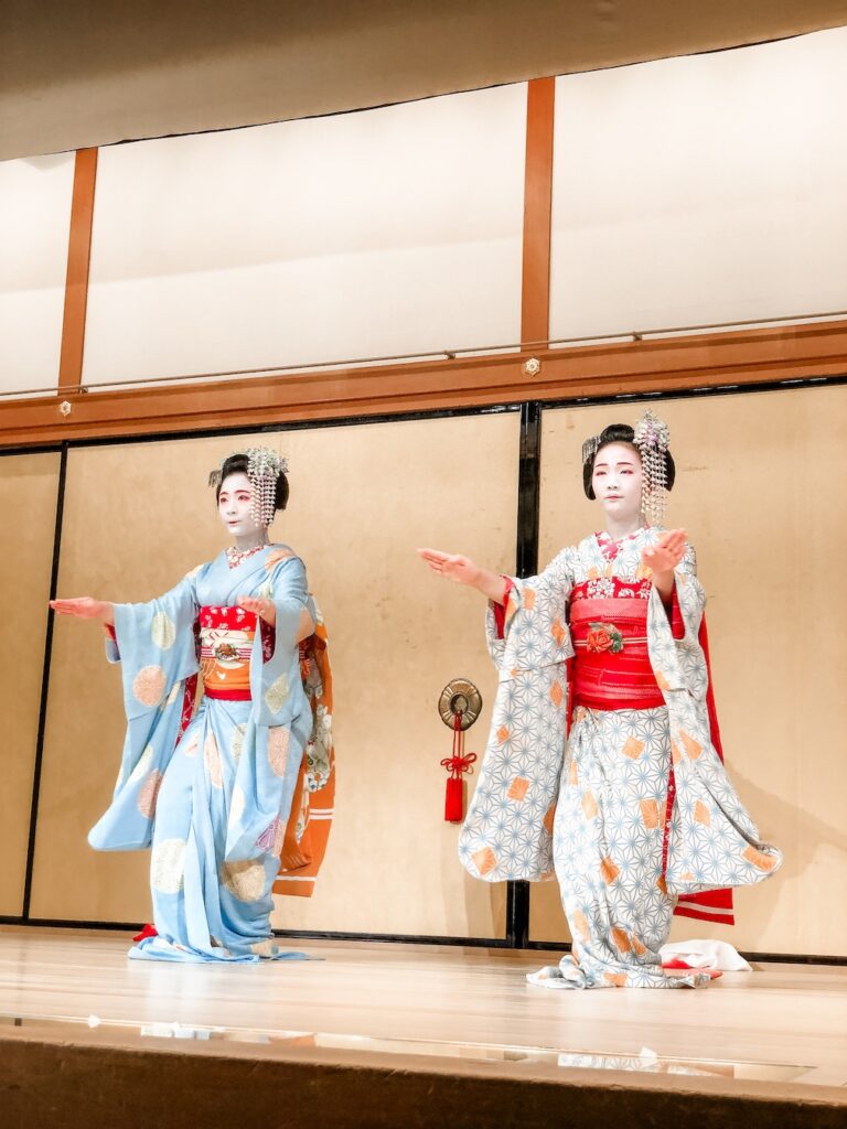 Two geisha perform a traditional dance at Gion Corner in Kyoto. One is dressed in a blue and red kimono, the other in white and red.