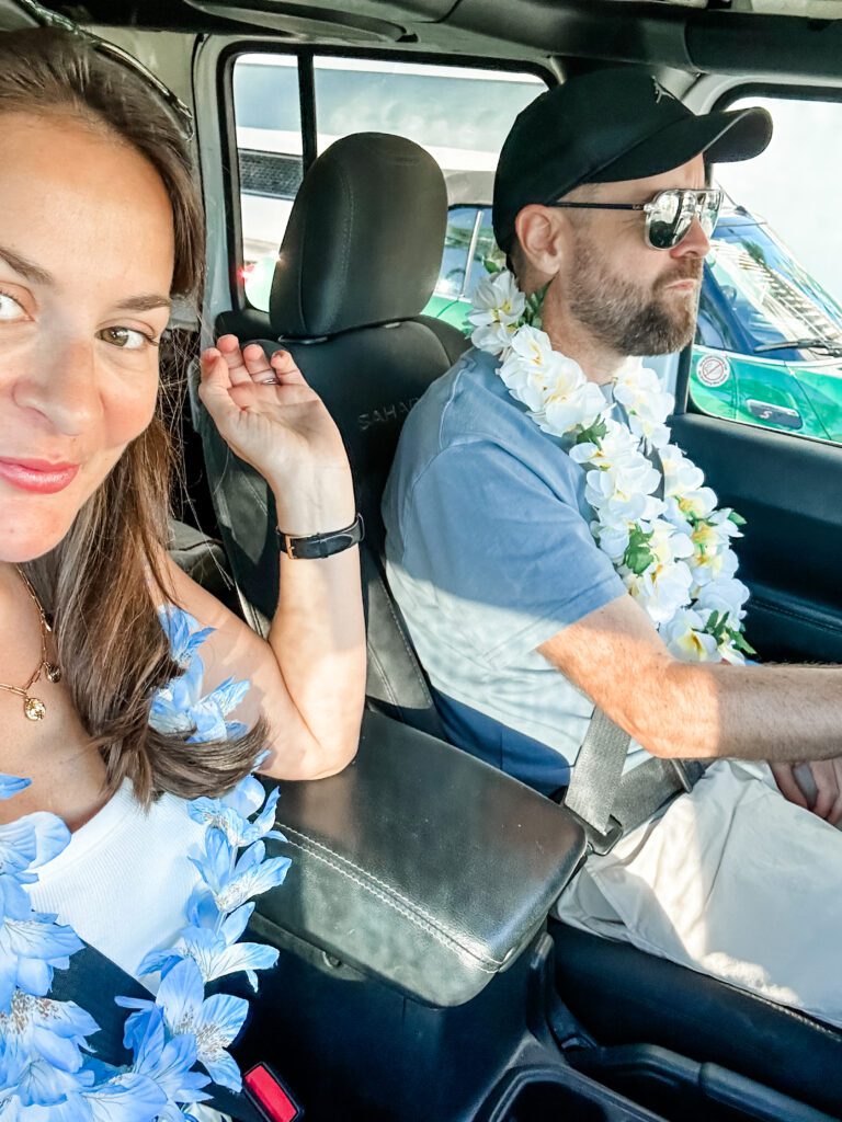 Rebecca and Chris Bowman, travel bloggers from Sydney, Australia, wearing floral leis and driving in a Jeep Wrangler for a day long road trip around the Hawaiian island of Oahu.