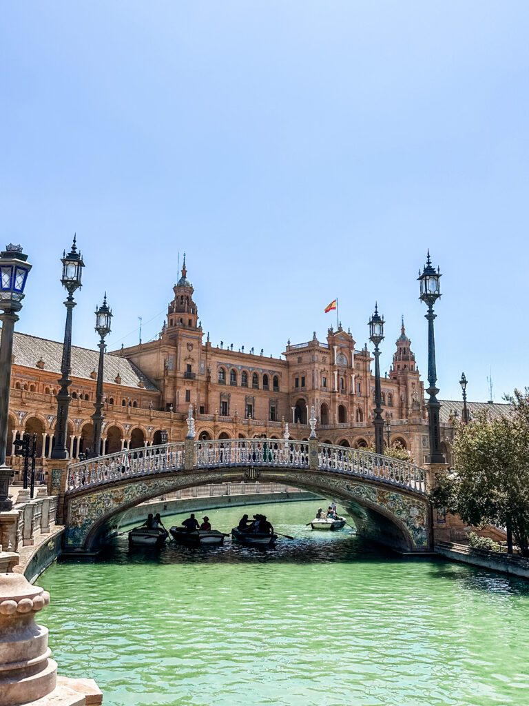 Alcázar in Seville on a hot summers day. The sky is blue and there are rowboats on the water in front of the Alcázar.
