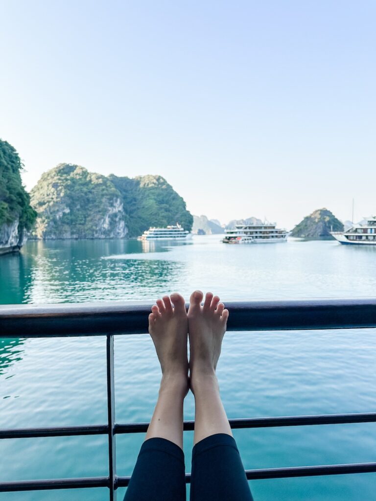 View of feet relaxing on the balcony of a Capella Cruise around Halong Bay. Other cruise ships are visible in the distance, before the limestone cliffs and emerald green waters. 
