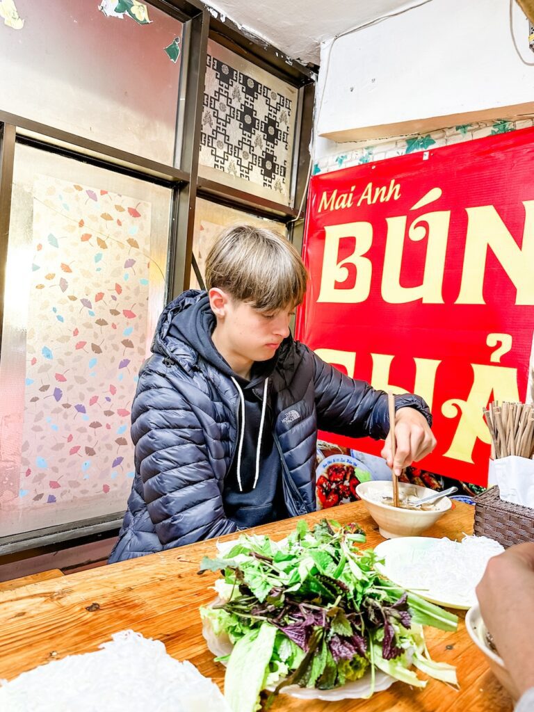 A teenage boy tucks into a bowl of Bun Cha in Hanoi. There is a bright red sign for the restaurant behind him and a huge bowl of green salad on the table.