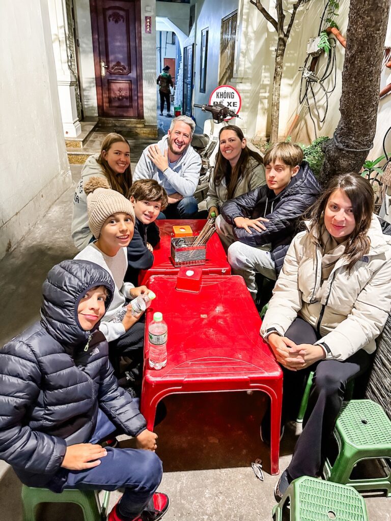A group of travellers sit around short tables on a side street in Hanoi waiting for their first taste of street food. The tables are red plastic. They are all wrapped up in winter coats and hats. 