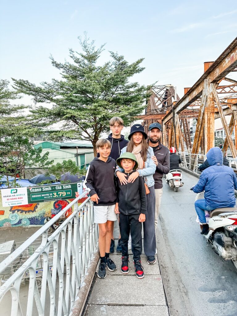 A family standing on the Long Bien Bridge in Hanoi at rush hour, with lots of mopeds driving on the street nearby.
