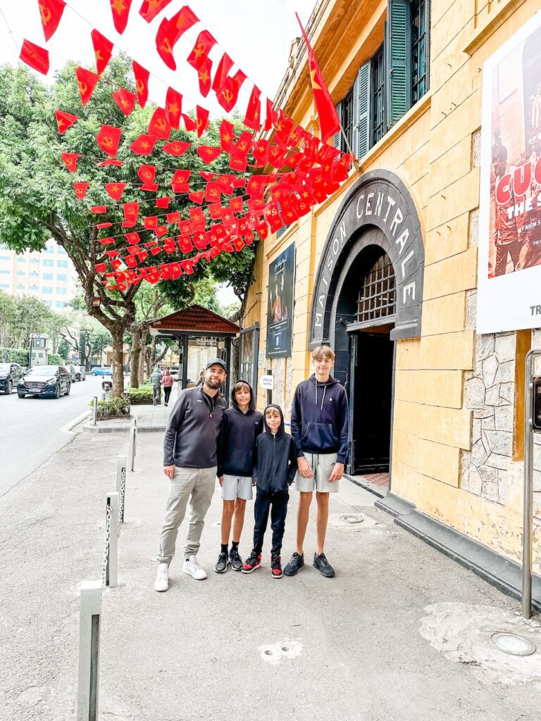 A photo of a family taken outside Maison Centrale in Hanoi. The walls of the building are yellow with green shutters and there are bright red Vietnam flags flying outside 