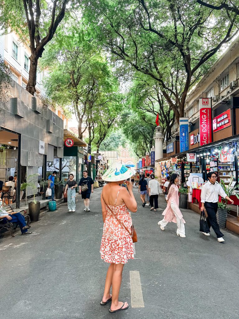 A woman stands looking along Saigon book street wearing a sun dress and traditional conical Vietnamese hat. The street is busy around her and sheltered by bright green trees.