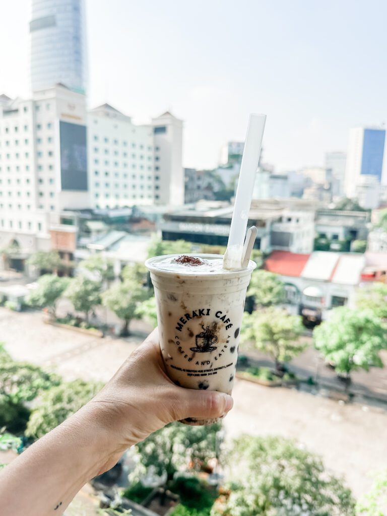 A photo of a woman's hand holding an iced coffee drink at Meraki Cafe in Ho Chi Minh City. The city is visible from the balcony, with both low lying and high rise buildings. 