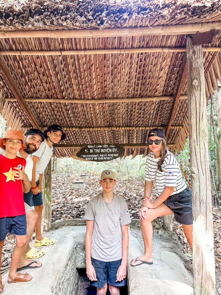 A family poses beside an opening to the Cu Chi Tunnels in Vietnam. One child wears a bright red t shirt with the Vietnamese star on the front. 