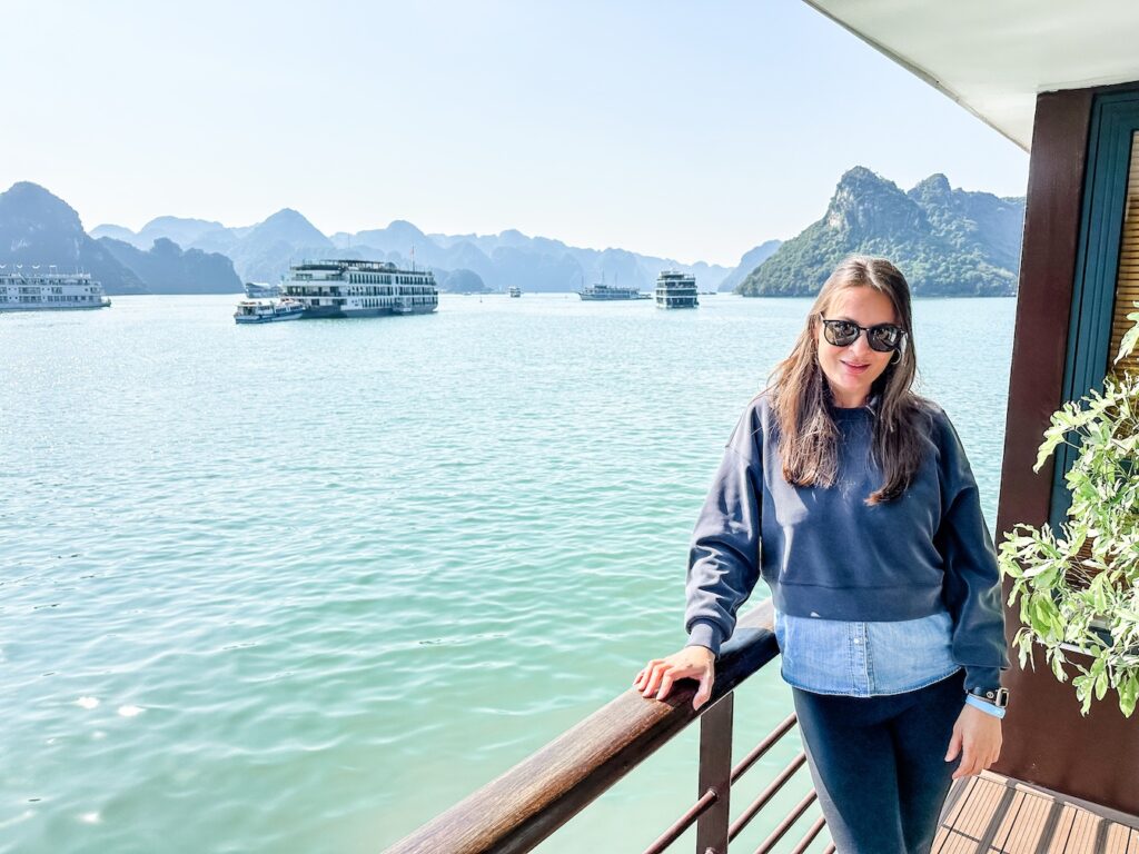 A woman stands on the balcony on the Capella Cruise around Ha Long Bay in Vietnam. The water is emerald green and the limestone cliffs behind are large and dramatic. 
